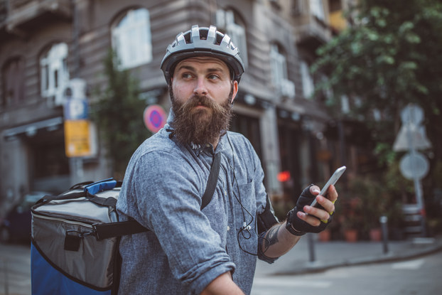 A bearded man in a city street intersection looks off-camera. He is carrying a cellphone and wearing a helmet and a large square backpack for carrying food. The photo is shot from the waist up, but the man is presumably sitting on a bike.
