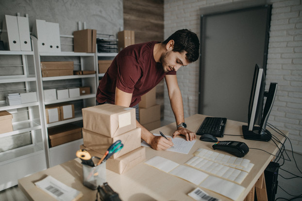A man stands over a table in a storage room. He writes something on a piece of paper. On the table around him are packages, a desktop monitor and keyboard, a credit card reader, and various receipts.