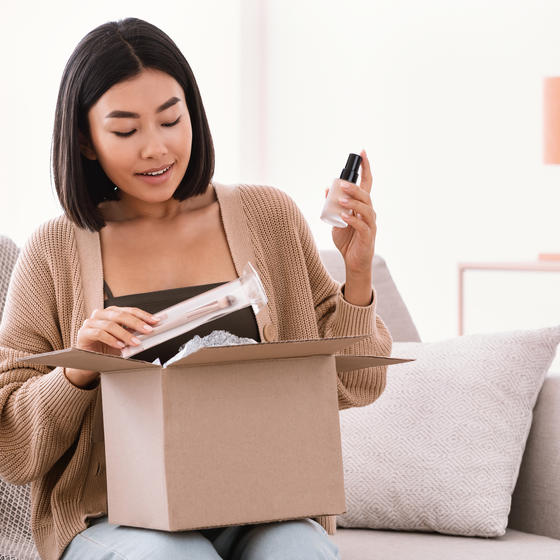 A woman sits on a couch and removes beauty products from a cardboard box.