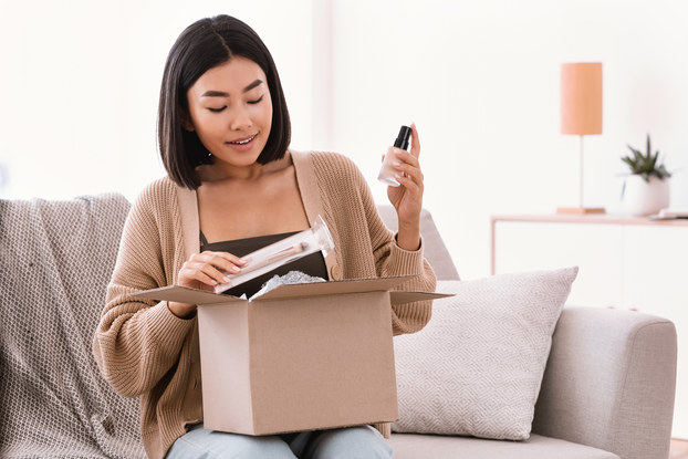 A woman sits on a couch and removes beauty products from a cardboard box.