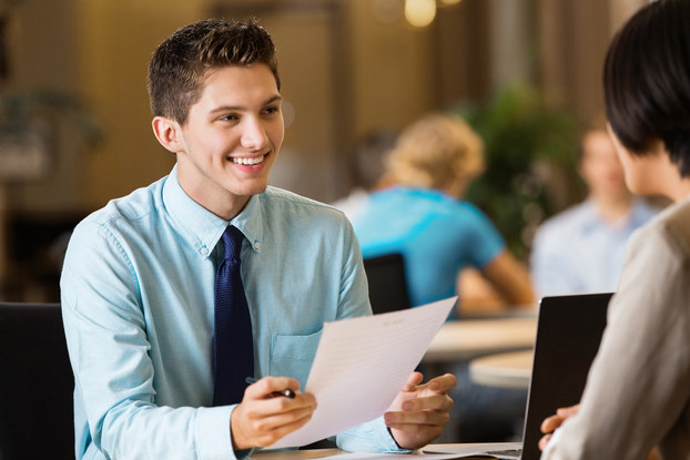 A young man in a shirt and tie sits across from an interviewer and holds a piece of paper.