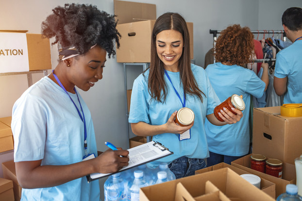 A group of people wearing light blue shirts and lanyards are boxing donations. One volunteer makes notes on a clipboard while another holds soup cans to be boxed. In the background, two more people look through a rack of clothes.
