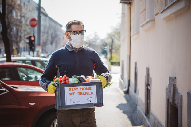 A man wearing a face mask and rubber gloves carries a box of vegetables away from a line of parked cars. A sign on the box reads "STAY HOME. WE DELIVER."