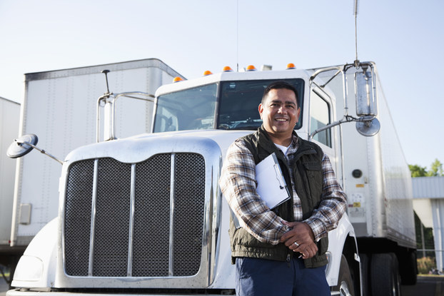 A man with a clipboard under one arm stands smiling in front of the cab of an 18-wheel truck.