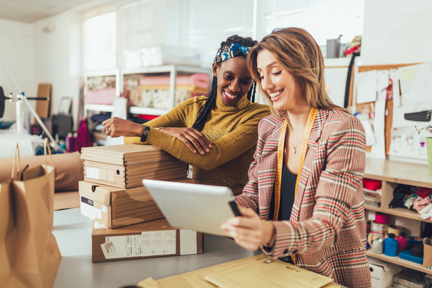 Two woman look at the screen of a tablet. The woman on the left leans on a pile of shoe boxes stacked on a worktable. In the background are shelves of fabric and bulletin boards covered in papers.