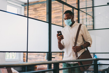  Man walking through office wearing a mask and looking at his phone. 
