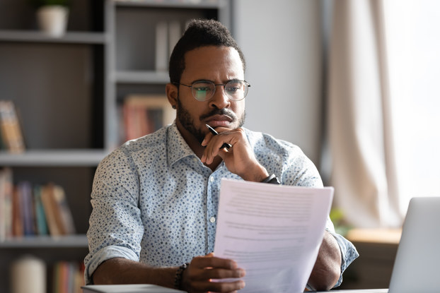 A man in glasses and a button-up shirt considers some paperwork with one hand on his chin. The hand on his chin also holds a pen.