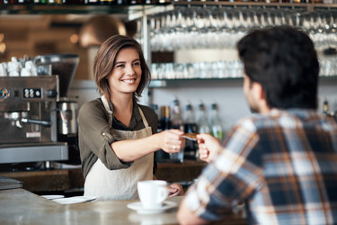  A barista hands a credit card back to a customer. A chrome brewing machine and various bottles of syrup stand behind the barista and a white mug on a saucer is placed on the countertop in front of her. 