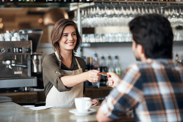 A barista hands a credit card back to a customer. A chrome brewing machine and various bottles of syrup stand behind the barista and a white mug on a saucer is placed on the countertop in front of her.