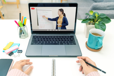  A POV shot of someone watching a class on a laptop and taking notes in a notebook. The laptop screen shows a woman gesturing to an easel displaying a large pad of paper covered with notes. 