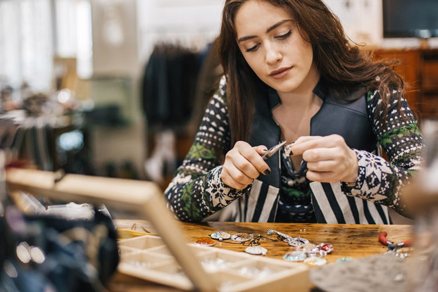 A woman sits at a table and uses a set of pliers to adjust a small piece of jewelry. Other pieces of jewelry -- namely pendants with various designs -- lay on the table before her, along with an open wooden box with several small compartments.