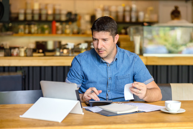 A man sits at a wooden counter and types on a calculator while looking at the screen of a tablet. In one hand, he holds a receipt and a folder of papers sits on the counter in front of him.