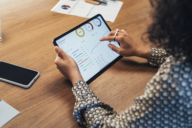  An over-the-shoulder shot of a woman examining charts and data on the screen of a tablet. 
