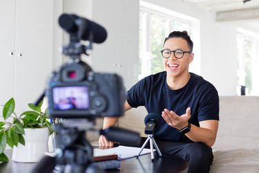  A male vlogger sits on the edge of a couch and speaks to a camera. The camera, which has a microphone attachment on top of it, stands on a tripod in the foreground, out of focus. 