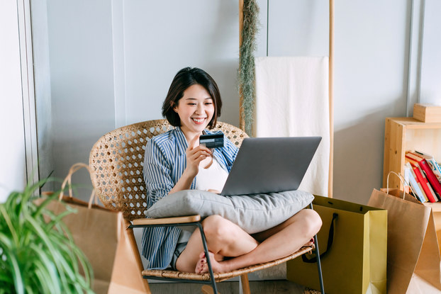 Woman shopping on laptop holding credit card while sitting on a rattan chair in her living room.