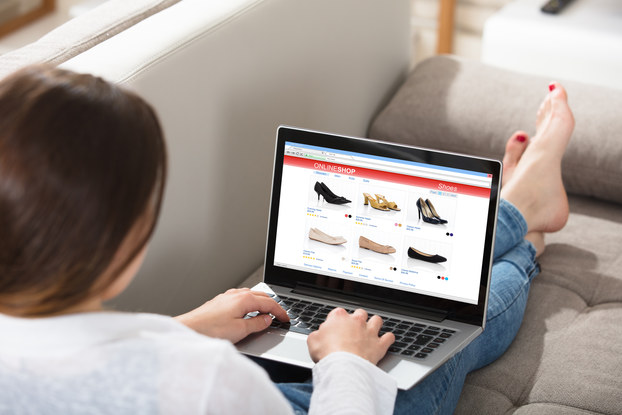 An over-the-shoulder shot of a woman browsing an online shoe store on her laptop. She reclines on a couch with her legs stretched out in front of her. Her feet are bare and her toenails are painted red.