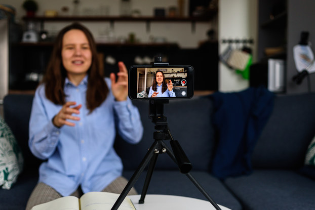 A smartphone positioned horizontally on a tripod films a woman speaking and gesturing. In the background, out of focus, is the woman herself.