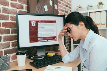  A woman sits at a desk with one hand to her forehead and her eyes closed. She looks defeated. The computer monitor on her desk displays a red and white window filled with text. An icon of a closed padlock suggests that the computer is locked. 