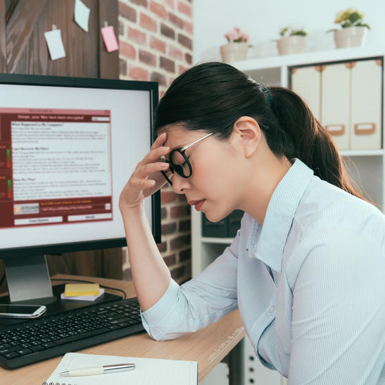 A woman sits at a desk with one hand to her forehead and her eyes closed. She looks defeated. The computer monitor on her desk displays a red and white window filled with text. An icon of a closed padlock suggests that the computer is locked.