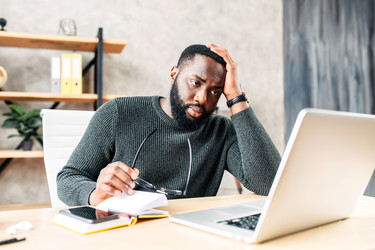  Frustrated man holds glasses and looks at laptop in an office. 