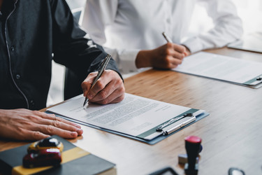  A below-the-neck shot of two people in button-up shirts sitting side by side at a table signing paperwork. The paperwork is attached to two clipboards. Also on the table but out-of-focus are a book and a rubber stamp. 