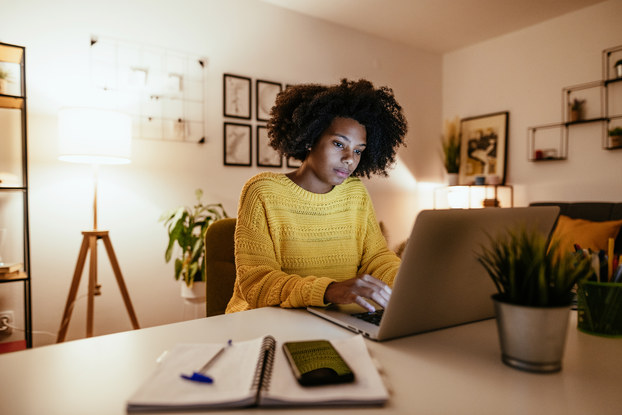 A young woman uses a laptop in a dimly lit modern living room. A notebook, pen, and smartphone are on the table next to her and light comes from a couple of lamps standing in the room behind her.