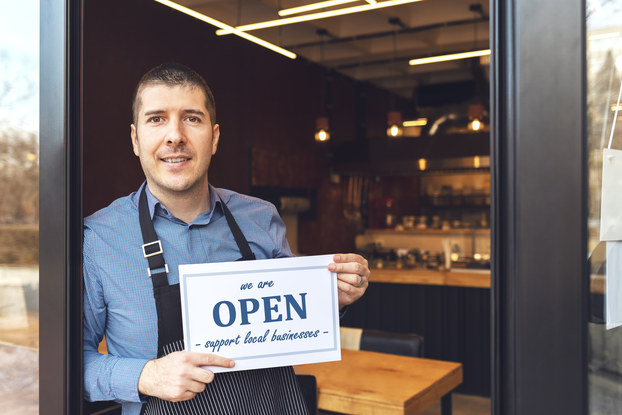A man wearing an apron stands in the entry way of his business. In the background, the store is outfitted with wood-top tables and shelves of dark bottles. The man holds a sign that says "we are OPEN" and "supporter small businesses." "OPEN" is written in bold blue letters, with the other words arranged above and below in blue cursive.
