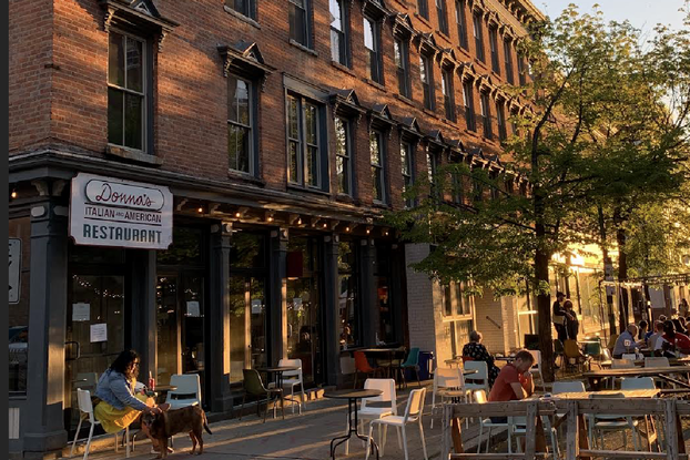 Outdoor seating outside a restaurant hosts customers with a tree and brick building in the background.