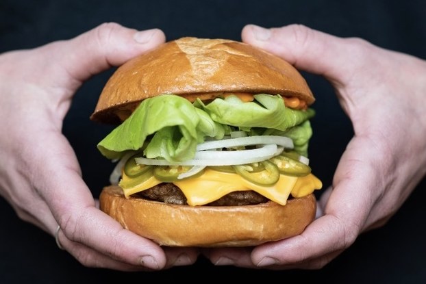 A tall cheeseburger with toppings is held in front of a black background.