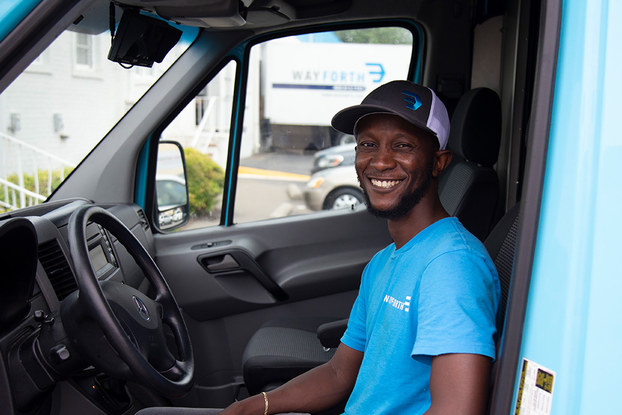 A smiling man wearing a cap sits in the driver's seat of a vehicle.