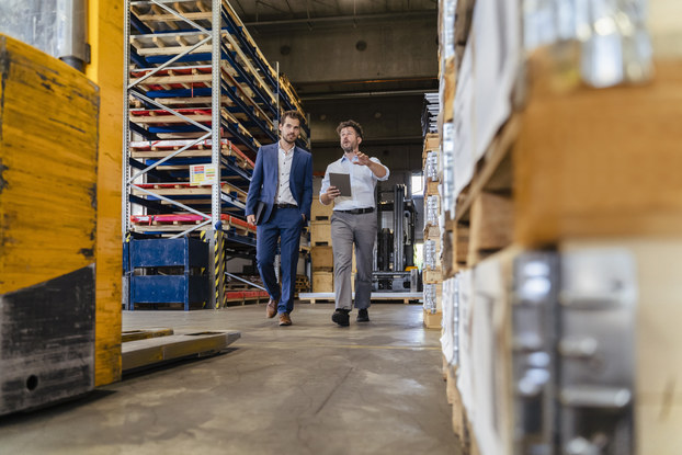 A wide shot of a pair of men walking through an aisle of a large warehouse. The man on the right is holding a clipboard and speaking to the man on the left, who wears a business suit. Behind them are large metal shelves holding wooden pallets. In the foreground, near the camera, are a forklift and a pile of pallets holding shrink-wrapped goods.