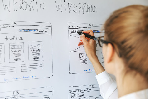 A woman seen from behind adds to a drawing on a whiteboard. The drawings show various pages of a website and their layouts under the heading "WEBSITE WIREFRAME."