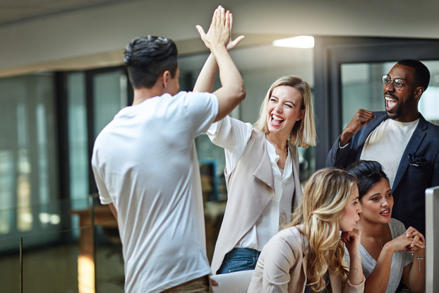 Several employees are in an office. Two of them are high-fiving and celebrating something. A third employee is also celebrating, standing near them and pumping his fist, his mouth open in a cheer. Two other employees are sitting at a computer in front of them, discussing something and not reacting to the people behind them.