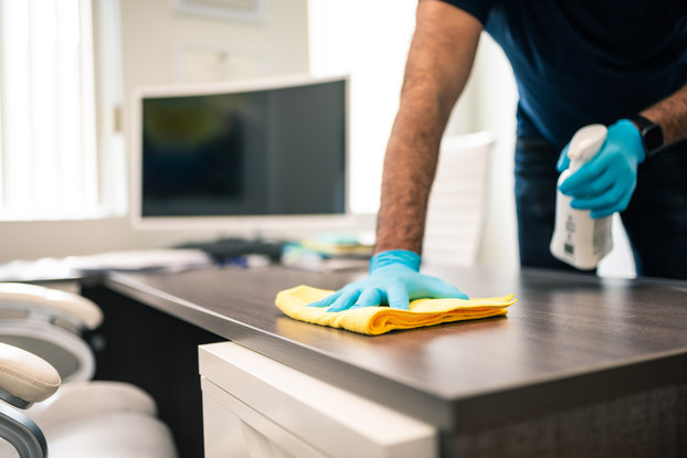 A person wearing light blue latex gloves uses a spray bottle and a yellow cloth to clean the top of a desk. The person's head and shoulders are out of frame. A room with large windows and a wide-screen TV can be seen out-of-focus in the background.