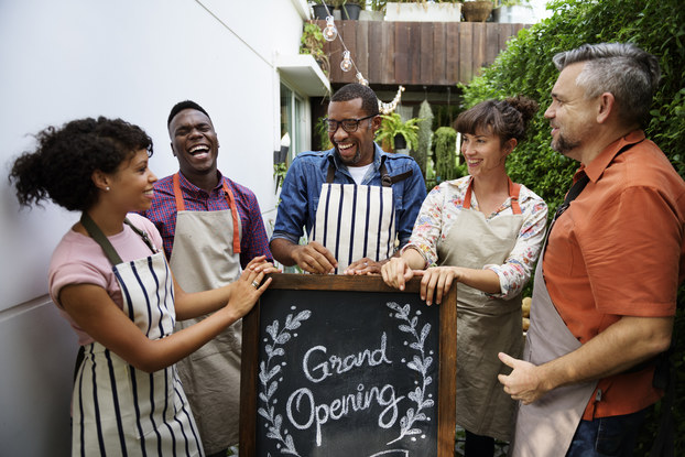 A group of five people wearing aprons stand around a chalkboard sign. The sign reads "Grand Opening" and the words are surrounded by a laurel wreath drawn in chalk. The five people are of different races and genders, and they stand with their hands on top of the sign, facing each other and smiling or laughing in celebration.