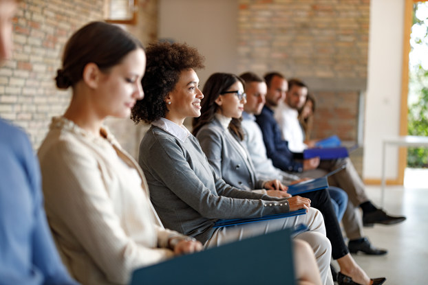 An angled side view of several job candidates dressed in business casual wear. The candidates are sitting in a row in a large room with brick walls. Most of the candidates are out of focus, but one woman in the middle is in-focus. She is looking up and straight-ahead at something out-of-frame, and she is smiling.