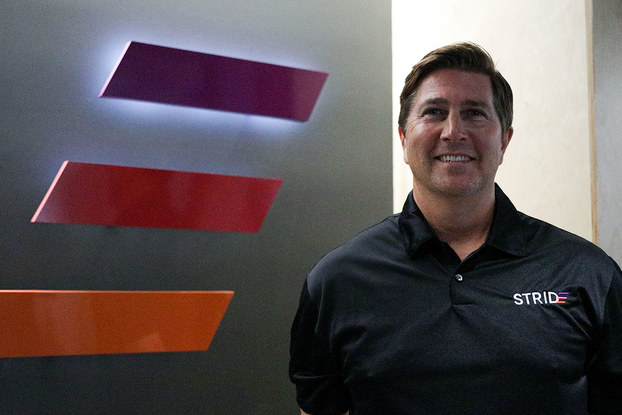 A shot of Jeff Stokes, president of STRIDE, standing next to the STRIDE logo (three parallel horizontal lines angled as if they're moving forward; the top line is purple, the middle red, and the bottom orange).