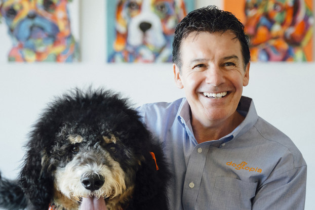 A headshot of Neil Gill, CEO of Dogtopia. He is sitting to the right of a large black-and-white dog wearing an orange bandanna and red collar.
