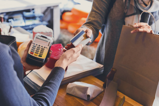 A close-up of a pair of two hands exchanging a credit card. The receiving hand belongs to someone holding a credit card reader. The giving hand belongs to someone with their other hand on a paper bag of merchandise.