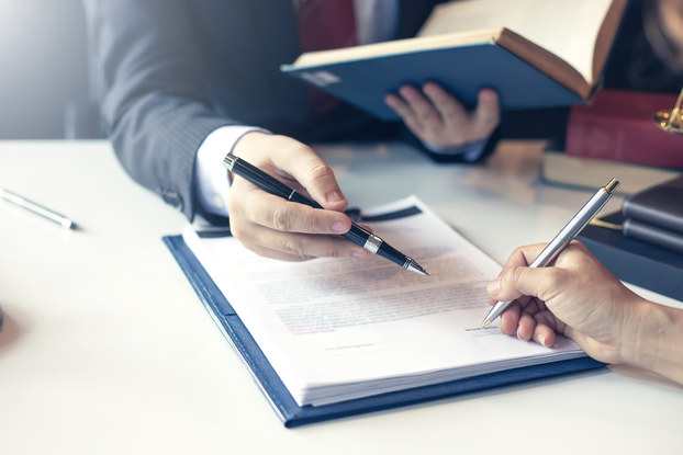 A close-up on the hands of two people sitting on either side of a white desk. One person's right hand signs the top paper of a stack of papers. The other person's right hand uses a pen to gesture to what should be signed. Their other hand holds an open book.