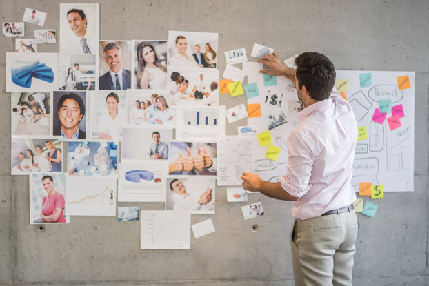 A man stands with his back to the camera in front of a gray concrete wall covers with photos of various people and objects and a large handwritten flowchart adorned with blue, orange, yellow and pink sticky notes.