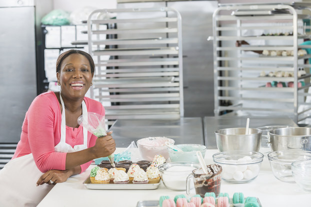 A Black woman wearing a pink shirt and a white apron leans against a counter in a large kitchen. On the counter beside her are mixing bowls, some filled with whipped icing, and trays of cupcakes iced with mint green, pastel pink, white and brown icing. An icing bag is in the woman's hand. In the background are cooling racks and industrial chrome refrigerators.