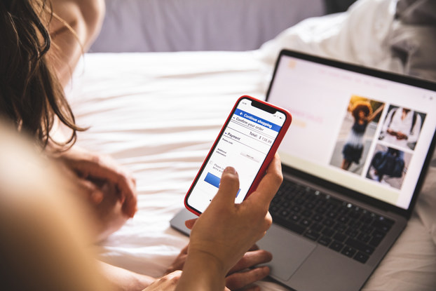 An over-the-shoulder shot of a person sitting on a bed and using a smartphone to view a website. In the background, out of focus, a laptop showing a different website sits on top of the white sheet covering the bed.