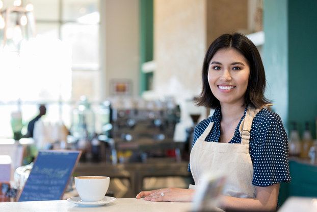 A young woman wearing a khaki apron stands behind a counter at a coffee shop. A large white cup of coffee sits on a saucer on the counter in front of her. In the background, out of focus, are a small chalkboard with chalk writing on it and various implements used for brewing and serving coffee.