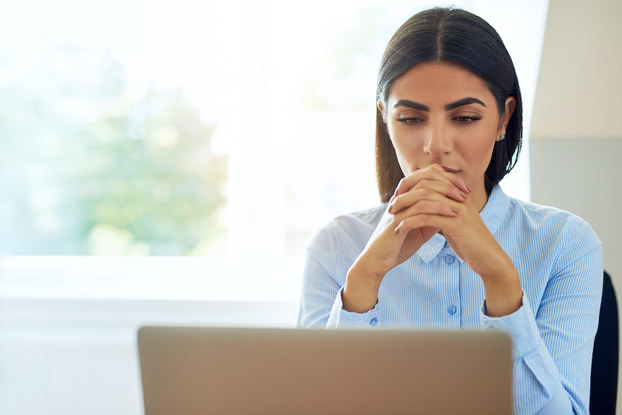 Serious young businesswoman with a worried expression sitting reading information on her laptop with clasped hands.