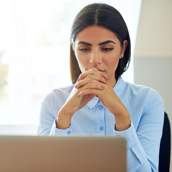 Serious young businesswoman with a worried expression sitting reading information on her laptop with clasped hands.
