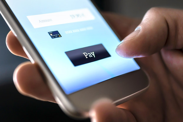 A close-up on a hand holding a smartphone. The thumb hovers over a button reading "Pay" on the phone's screen.