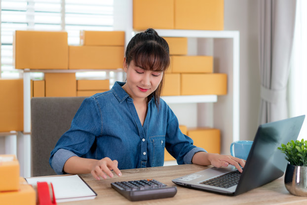 A woman sits at a wooden desk and uses a calculator with her right hand. Her other hand rests on a laptop. In the background are shelves of boxes.