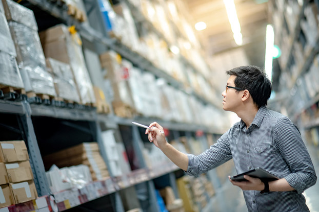 A young man in a button-up shirt stands in a cavernous warehouse, looking up at one of many industrial shelves holding wrapped boxes on wooden pallets. He holds an electronic tablet in one hand and uses the other hand to point a pen at the boxes, as if counting them.