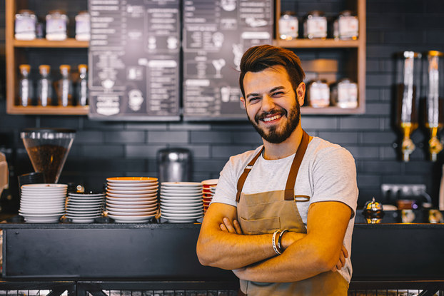 A man wearing a khaki apron stands smiling with his arms folded in front of a counter at a cafe and menu board at a cafe. The menu board is white text on a black background and is mounted on a wall between shelves of jars and carafes. The counter is stacked with plates of various sizes.